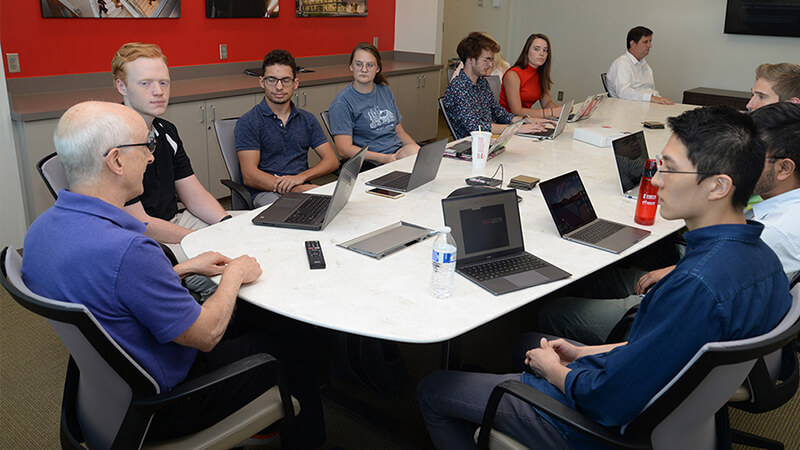 Students with laptops seated at large table listen to speaker seated at the head of the table.